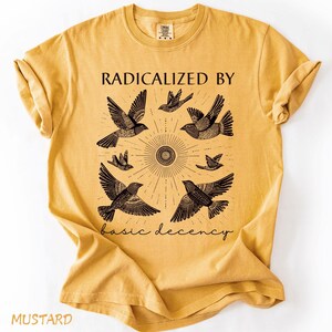 Puede incluir: Camiseta de color mostaza con un gráfico de pájaros estilizados que rodean un diseño de sol. El texto "RADICALIZED BY basic decency" está impreso en negro. La camiseta tiene mangas cortas y cuello redondo.