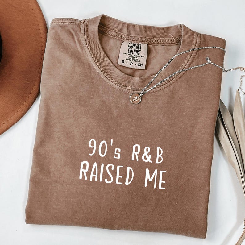 Puede incluir: Una camiseta marr&oacute;n con el texto "90's R&B RAISED ME" en blanco. La camiseta tiene un aspecto desgastado y est&aacute; doblada por la mitad.