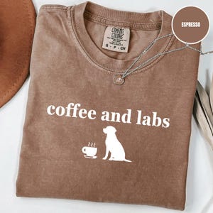 May include: Espresso-coloured t-shirt with the words "coffee and labs" in white. A white graphic of a coffee cup and a Labrador Retriever dog is below the text. The shirt has a crew neck and a small tag.