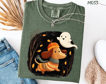 May include: A moss green Comfort Colors t-shirt featuring a whimsical illustration of a dachshund wearing a hat and sweater, with a friendly ghost. The design includes autumn leaves and trees, perfect for fall. A silver necklace is draped across the shirt.