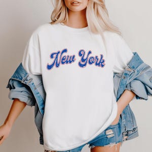 May include: White t-shirt with a blue and red "New York" graphic in a retro font.