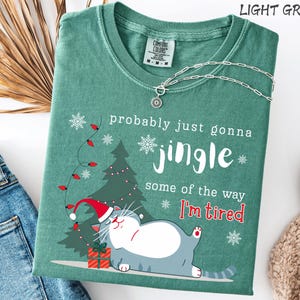 Puede incluir: Camiseta verde claro con el texto "probably just gonna jingle some of the way I'm tired". La camiseta presenta un gato de dibujos animados con un gorro de Pap&aacute; Noel, un &aacute;rbol de Navidad y copos de nieve. Un collar plateado est&aacute; drapeado sobre la camiseta.