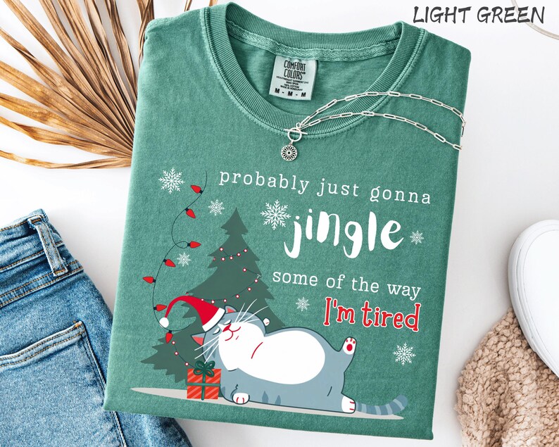 Puede incluir: Camiseta verde claro con el texto "probably just gonna jingle some of the way I'm tired". La camiseta presenta un gato de dibujos animados con un gorro de Pap&aacute; Noel, un &aacute;rbol de Navidad y copos de nieve. Un collar plateado est&aacute; drapeado sobre la camiseta.