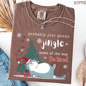 Puede incluir: Camiseta color espresso con un gr&aacute;fico tem&aacute;tico navide&ntilde;o. El dise&ntilde;o presenta un gato con gorro de Pap&aacute; Noel, un &aacute;rbol de Navidad, copos de nieve y el texto "probably just gonna jingle some of the way I'm tired."