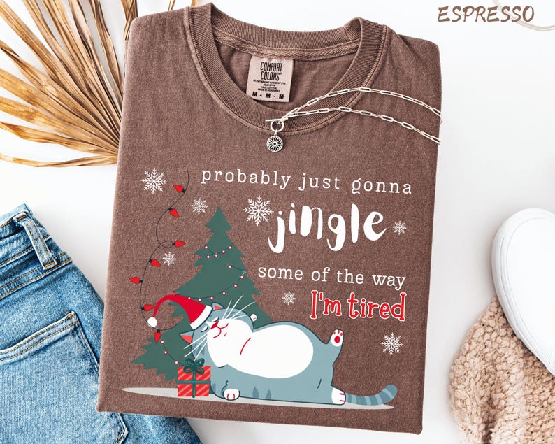Puede incluir: Camiseta color espresso con un gr&aacute;fico tem&aacute;tico navide&ntilde;o. El dise&ntilde;o presenta un gato con gorro de Pap&aacute; Noel, un &aacute;rbol de Navidad, copos de nieve y el texto "probably just gonna jingle some of the way I'm tired."