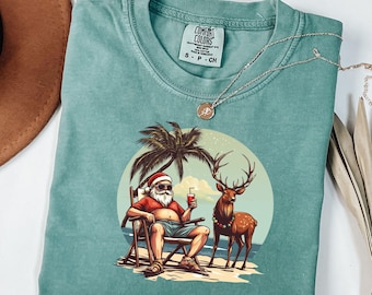 Könnte beinhalten: Ein türkisfarbenes Comfort Colors T-Shirt mit einer Grafik von Santa Claus, der sich auf einem Strandstuhl mit einem Getränk, einer Palme und einem Rentier entspannt. Das T-Shirt hat einen Rundhalsausschnitt und kurze Ärmel. Eine silberne Halskette mit Anhänger ist auf dem T-Shirt drapiert.