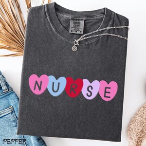 May include: Dark gray t-shirt with the word "NURSE" spelled out in colorful heart-shaped letters. A silver necklace with a small pendant is draped across the shirt. The shirt is made of a soft material.