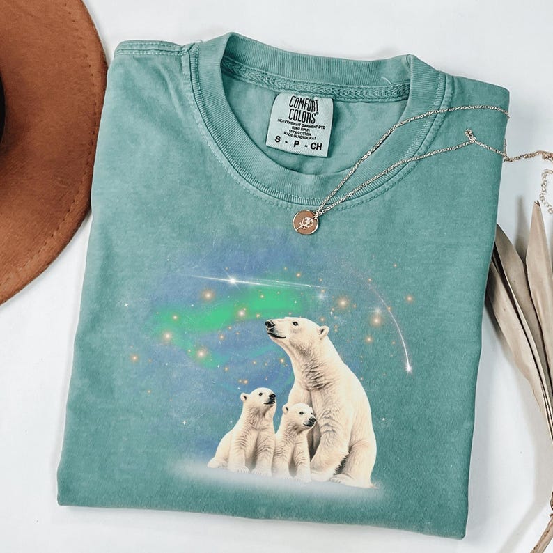 May include: A teal blue t-shirt with a graphic of three polar bears looking up at the aurora borealis. The shirt has a tag that reads "Comfort Colors" and "Heather Garment Dye".