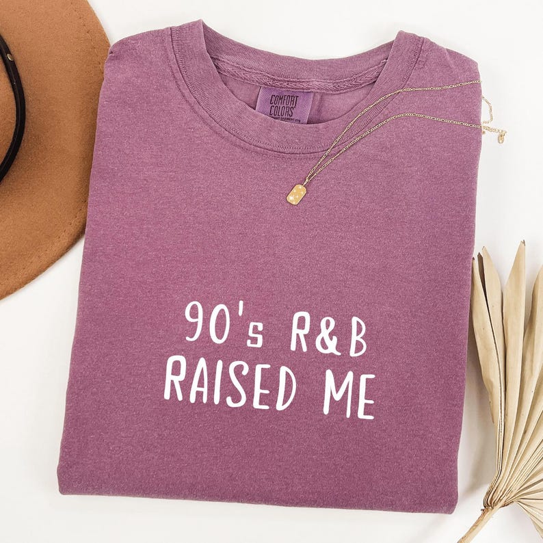 Puede incluir: Una camiseta de color malva con el texto "90's R&B RAISED ME" en letras blancas.