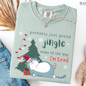 Puede incluir: Camiseta verde claro con el texto "probably just gonna jingle some of the way I'm tired". El dise&ntilde;o presenta un gato de dibujos animados con un gorro de Pap&aacute; Noel, un &aacute;rbol de Navidad, copos de nieve y un regalo. Un collar plateado est&aacute; sobre la camiseta.
