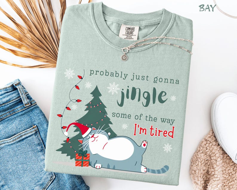 Puede incluir: Camiseta verde claro con el texto "probably just gonna jingle some of the way I'm tired". El dise&ntilde;o presenta un gato de dibujos animados con un gorro de Pap&aacute; Noel, un &aacute;rbol de Navidad, copos de nieve y un regalo. Un collar plateado est&aacute; sobre la camiseta.