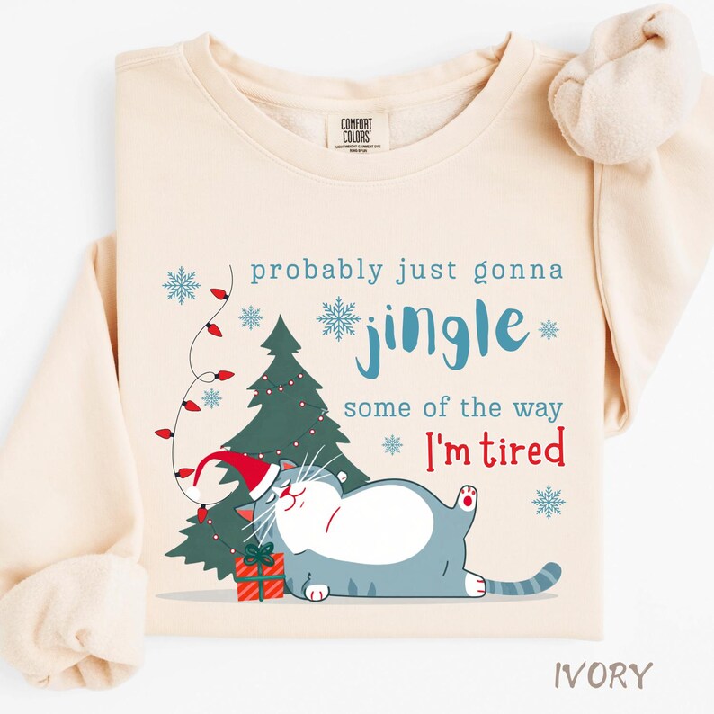 Puede incluir: Sudadera de color marfil con un gr&aacute;fico de gato navide&ntilde;o. El dise&ntilde;o incluye un gato con gorro de Pap&aacute; Noel, un &aacute;rbol de Navidad y el texto "probably just gonna jingle some of the way, I'm tired".