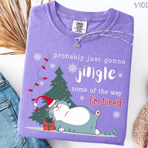 Puede incluir: Camiseta violeta Comfort Colors con un gr&aacute;fico de gato navide&ntilde;o. El dise&ntilde;o incluye un &aacute;rbol de Navidad, copos de nieve y el texto "probably just gonna jingle some of the way I'm tired". Un collar plateado est&aacute; drapeado sobre la camisa.
