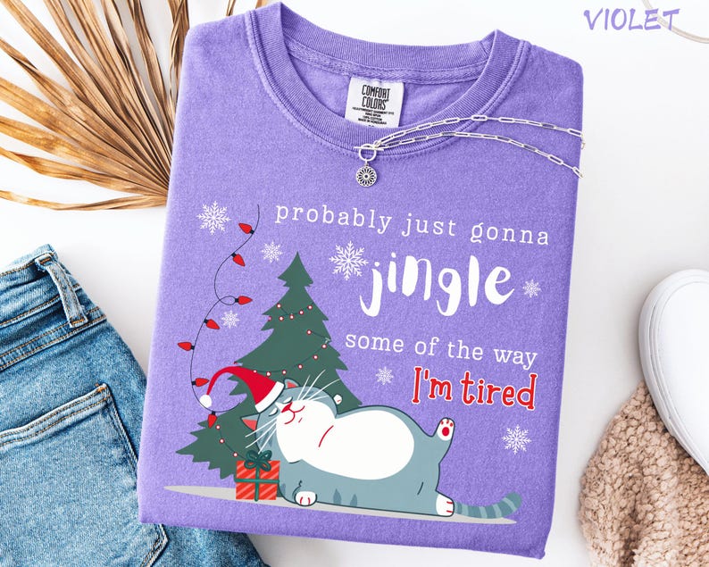 Puede incluir: Camiseta violeta Comfort Colors con un gr&aacute;fico de gato navide&ntilde;o. El dise&ntilde;o incluye un &aacute;rbol de Navidad, copos de nieve y el texto "probably just gonna jingle some of the way I'm tired". Un collar plateado est&aacute; drapeado sobre la camisa.