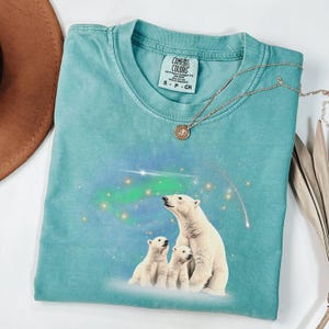 May include: A teal blue t-shirt with a graphic of three polar bears looking up at the aurora borealis. The shirt has the text "Comfort Colors" and "S - P - CH" on the tag.