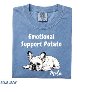 May include: A blue jean colored t-shirt featuring a white graphic of a French Bulldog lying down. The text "Emotional Support Potato" is printed in white above the dog, and the name "Mila" is printed in white script below the dog illustration.