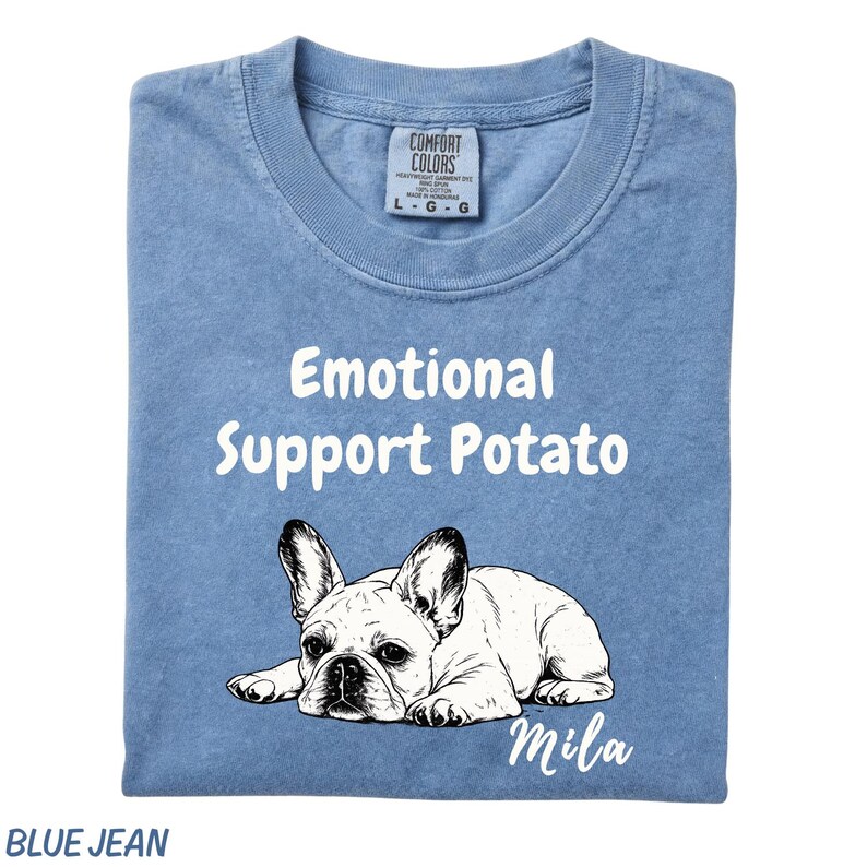 May include: A blue jean colored t-shirt featuring a white graphic of a French Bulldog lying down. The text "Emotional Support Potato" is printed in white above the dog, and the name "Mila" is printed in white script below the dog illustration.