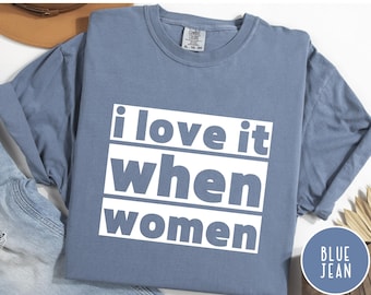 Op de afbeelding: Blauw spijkerbroekkleurig T-shirt met de tekst "i love it when women" in een witte, vette letter. Het shirt heeft een effen kleur en een ronde hals. Het shirt is opgevouwen en ligt op een wit oppervlak.