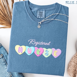 Op de afbeelding: Blauwe jeanskleurige t-shirt met het woord "Registered" in cursief boven vijf kleurrijke hartvormige ontwerpen die "NURSE" spellen. Een zilveren ketting met een hanger ligt op de shirt. Het shirt is op een wit oppervlak gevouwen.