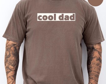 May include: A brown t-shirt with the words "cool dad" in a white rectangle. The shirt has short sleeves and a crew neck. A brown circle with the word "ESPRESSO" is also visible in the image.
