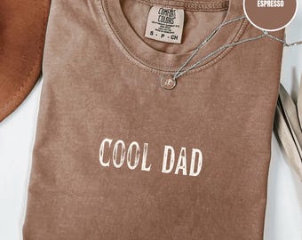 May include: Espresso-coloured t-shirt with the words "COOL DAD" printed in white. The shirt has a crew neck and a small tag at the neckline. A silver necklace with a pendant is draped across the shirt.