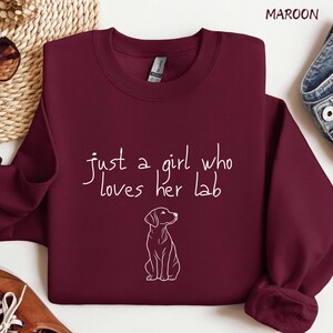 Puede incluir: Sudadera burdeos con la frase "just a girl who loves her lab" en escritura blanca. Un dibujo lineal blanco de un Labrador Retriever est&aacute; debajo del texto. La sudadera est&aacute; doblada.