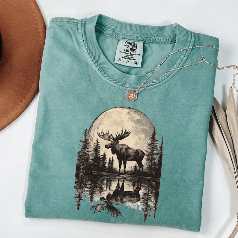 May include: A teal blue t-shirt with a graphic of a moose standing in front of a full moon and a reflection of the moose in a lake. The text "COMFORT COLORS" is on a white tag attached to the shirt.