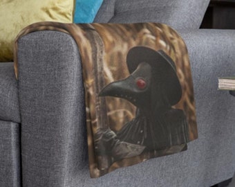 Può includere: Un divano grigio con una coperta che presenta un'illustrazione in bianco e nero di un medico della peste con un lungo becco e un cappello. La coperta è drappeggiata sul braccio del divano.