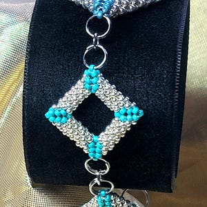 Può includere: Un braccialetto di perline con un design geometrico. Il braccialetto presenta perline argentate e turchesi disposte a forma di diamante. Il braccialetto è collegato con anelli di salto argentati ed è esposto su una superficie nera.