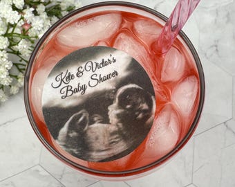 May include: A glass of pink liquid with ice cubes and a pink swirled straw. A circular edible image with the text "Kate & Victor's Baby Shower" and a grayscale sonogram image is in the drink.