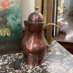 May include: A copper coffee pot with a brass lid and a handle. The pot has a rounded body and a slightly flared spout. The pot is sitting on a marble surface.