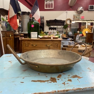 May include: A vintage, round, brass-toned pan with two curved handles. The pan sits on a distressed, light blue wooden table. The pan's interior shows signs of wear, suggesting age and use. The background includes French flags and antique furniture.