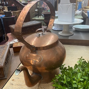 May include: A vintage copper kettle with a large, arched handle and a spout. The kettle has a rounded body and a lid with two small loops. The copper has a warm, reddish-brown hue, suggesting age and use. The kettle is likely for boiling water.