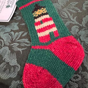May include: A hand-knitted Christmas stocking in green and red, featuring a lighthouse design. The lighthouse is striped in red and white, with a black and yellow top. A tag with text is attached.