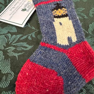 May include: A small, hand-knitted Christmas stocking featuring a lighthouse design. The stocking is primarily blue with a red toe and cuff, and a white lighthouse with black and yellow accents. The tag reads "Christmas Cove Designs".