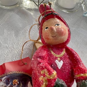 May include: A Christmas ornament featuring a whimsical figure in a red hooded outfit with gold accents and a white heart. The ornament has a painted face and a gold string for hanging. A tag with a Santa Claus illustration is attached.