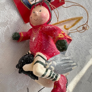 May include: A Christmas ornament featuring a whimsical figure in a red outfit with green accents, riding a gray and black bird. The ornament has white and gold wings and is suspended by a gold string. The figure has a smiling face and is decorated with glitter.