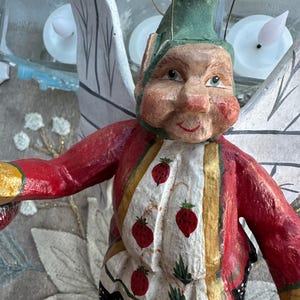 May include: A whimsical wooden fairy figurine with a green hat, red jacket, and white shirt adorned with red strawberry designs. The fairy has painted facial features and delicate, white-painted wings. The figurine is a decorative item.