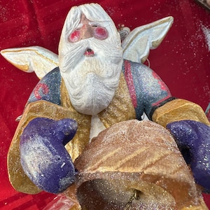 May include: A decorative Christmas angel figurine with a white beard, red cheeks, and gold-trimmed wings. The angel is wearing a gold and blue outfit and holding a bell. The figurine is covered in glitter.