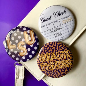 May include: Three circular pinback buttons. One features a white guest check design with the text "Guest Check" and "SERVING TIGER BAIT". Another is purple with white polka dots and gold "LSU" text. The third is purple and gold with "GEAUX TIGERS".