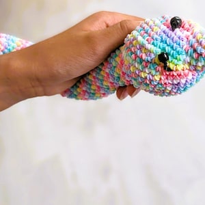 May include: A snake toy crafted from multicoloured rubber bands in pastel shades of pink, blue, yellow, and white. The snake features two small black beads for eyes and a protruding red tongue made of rubber bands, with a textured, woven surface.