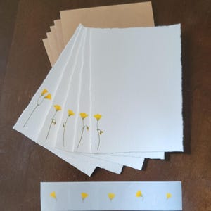 May include: A stack of white stationery with deckled edges, each adorned with a delicate yellow flower design. Kraft paper envelopes are stacked behind the stationery. A strip of stickers with yellow flower designs is at the bottom.