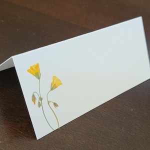 May include: A folded white place card with a floral design. The card features two yellow flowers with green stems and buds. The card is blank, offering space for personalized text.