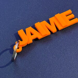 Può includere: Portachiavi in plastica arancione con il nome "JAME" in grassetto. Il portachiavi ha un anello in metallo argentato.