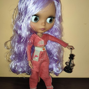 May include: A doll with long, wavy, lavender hair wearing a red jumpsuit with a mushroom patch and holding a small black lantern.