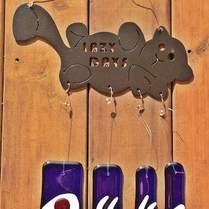 Könnte beinhalten: Ein Windspiel aus Metall in Form einer Katze mit den ausgeschnittenen Worten "Lazy Days". Das Windspiel hat vier hängende Glasstücke mit verschiedenen Designs in Blautönen, Weiß und Rot.