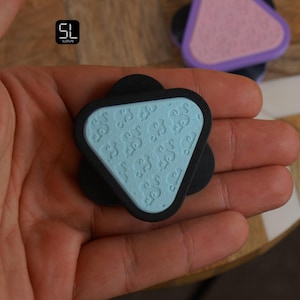 May include: A black phone grip with a light blue, triangular top. The light blue surface has a repeating, swirling pattern. Another grip with a pink top and purple border is visible in the background.