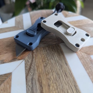 May include: Two compact utility knives with textured handles, one in slate blue and one in off-white. Each knife features a sliding mechanism, exposed metal blade, and four visible screws on the handle. A black cord is attached to the off-white knife.