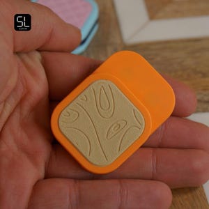 May include: Two orange, square-shaped objects with a beige wood grain design are held in a hand. The objects are slightly overlapping. The background is a wooden surface.