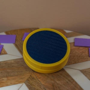 May include: A yellow and navy blue round object with a textured surface, surrounded by several small purple rectangular tiles with various patterns. The items are arranged on a wooden surface with a chevron pattern.
